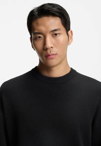 Schwarzer Strickpullover mit einem gerippten Rundhalsausschnitt und weicher Textur, der eine entspannte Passform und ein minimales Design bietet. Keine Muster oder sichtbaren Beschläge.