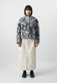 Fleece jacket with a black and white marbled pattern, high collar, and full zipper, paired with a cream-colored long skirt and black sneakers.