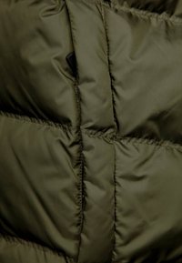 Giacca puffer in un profondo verde oliva, caratterizzata da sezioni trapuntate, tessuto in nylon liscio e una leggera lucentezza. Include un dettaglio con tasca nascosta.