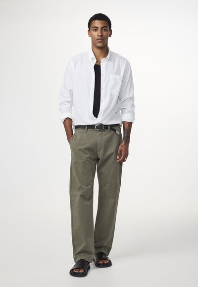Witte lange mouwen shirt over een zwart t-shirt, olijfgroen broek, zwarte riem en zwarte sandalen. Heeft een losse pasvorm en een voorzak.