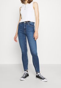 Top corto de tirantes blanco acanalado con dobladillo ondulado, combinado con jeans ajustados de mezclilla oscura y zapatillas altas negras con cordones blancos.