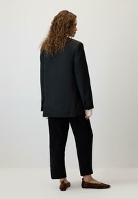 Melnā piegriezuma žakete ar strukturētām plecu daļām, apvienota ar melnajiem kordūras biksēm un brūnām leopardu raksta kurpēm; modelis skatīts no aizmugures.