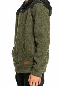 Sudadera color verde oliva con un cuerpo texturizado, capucha negra con un patrón, cierre frontal y un detalle de parche de cuero en el dobladillo. Cuenta con bolsillos laterales.