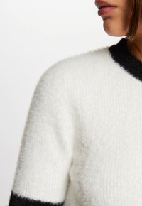 Suéter blanco esponjoso con un tejido texturizado, que presenta puños y cuello negro cortos. Muestra un tejido suave y cálido con detalles de color en contraste.