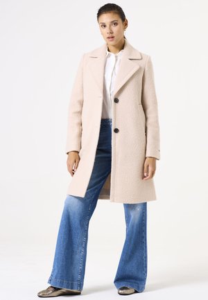Abrigo texturizado beige con cuello con muesca y tres botones negros; combinado con jeans de pierna ancha y zapatos metálicos.