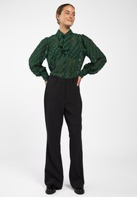 Groene blouse met patroon, een gerimpelde kraag, doorzichtig materiaal en lange ballonmouwen, gecombineerd met zwarte high-waisted broek en elegante schoenen.