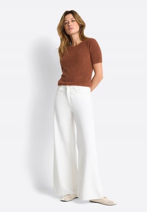 Kobieta ubrana w brązowy krótki sweter z krótkim rękawem, szerokie białe spodnie i kremowe wsuwane buty z plecionki, stojąca z rękami za plecami.