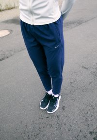 Granatowe spodnie sportowe o dopasowanym kroju, z elastycznym pasem i logo Nike, w połączeniu z czarnymi trampkami i białymi skarpetkami na powierzchni chodnika.