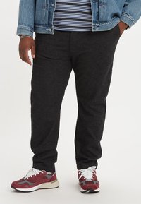 Zwarte tapse broek gemaakt van gestructureerde stof, gedragen met een gestreept shirt en spijkerjack, gecombineerd met rode en grijze sneakers.