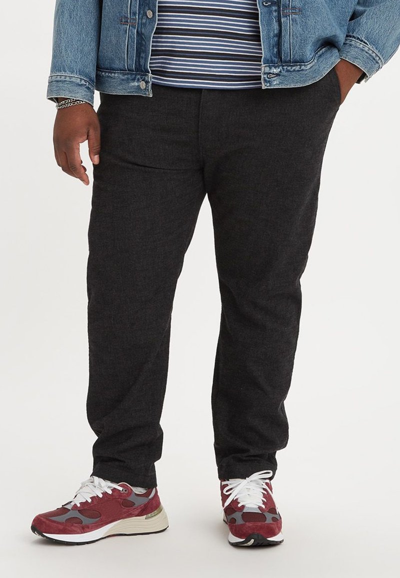Zwarte tapse broek gemaakt van gestructureerde stof, gedragen met een gestreept shirt en spijkerjack, gecombineerd met rode en grijze sneakers.