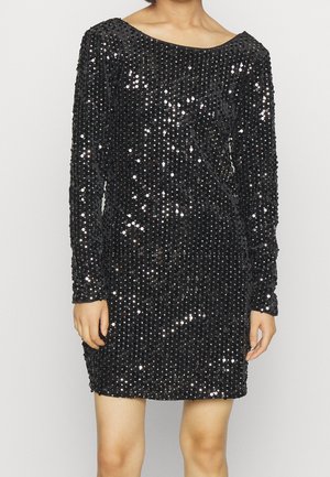 Femme portant une mini-robe noire à manches longues recouverte de petits sequins brillants sur un fond uni.