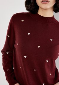Pull en maille bordeaux avec un col haut, orné de cœurs brodés éparpillés dans des tons métalliques. Texture douce, manches longues.