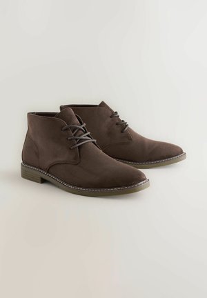 Botas de ante marrón de tobillo con cordones, con una punta ligeramente afilada y un tacón de madera bajo, exhibidas sobre un fondo claro.