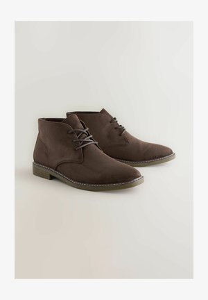 Braune Wildleder-Stiefeletten mit Schnürung, die eine leicht spitz zulaufende Schuhspitze und einen niedrigen Holzabsatz aufweisen, vor einem hellen Hintergrund präsentiert.