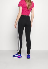 Černé teplákové kalhoty s bílými pruhy po stranách, doplněné růžovým krátkým topem a modrými sportovními botami s růžovými akcenty.