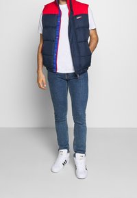 Tmavě modrá péřová vesta s červenou horní částí a modrým zipem, nošená přes bílé tričko. Kombinováno s modrými džínami a bílými teniskami.