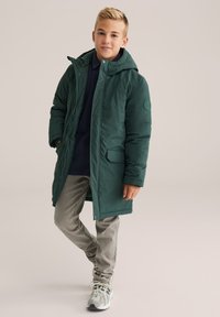 Jauns zēns stāv, ģērbies tumši zaļā kapuces ziemas mētelī, tumši zili krekls, bēšas bikses un pelēki sporta apavi, pret vienkāršu fonu.