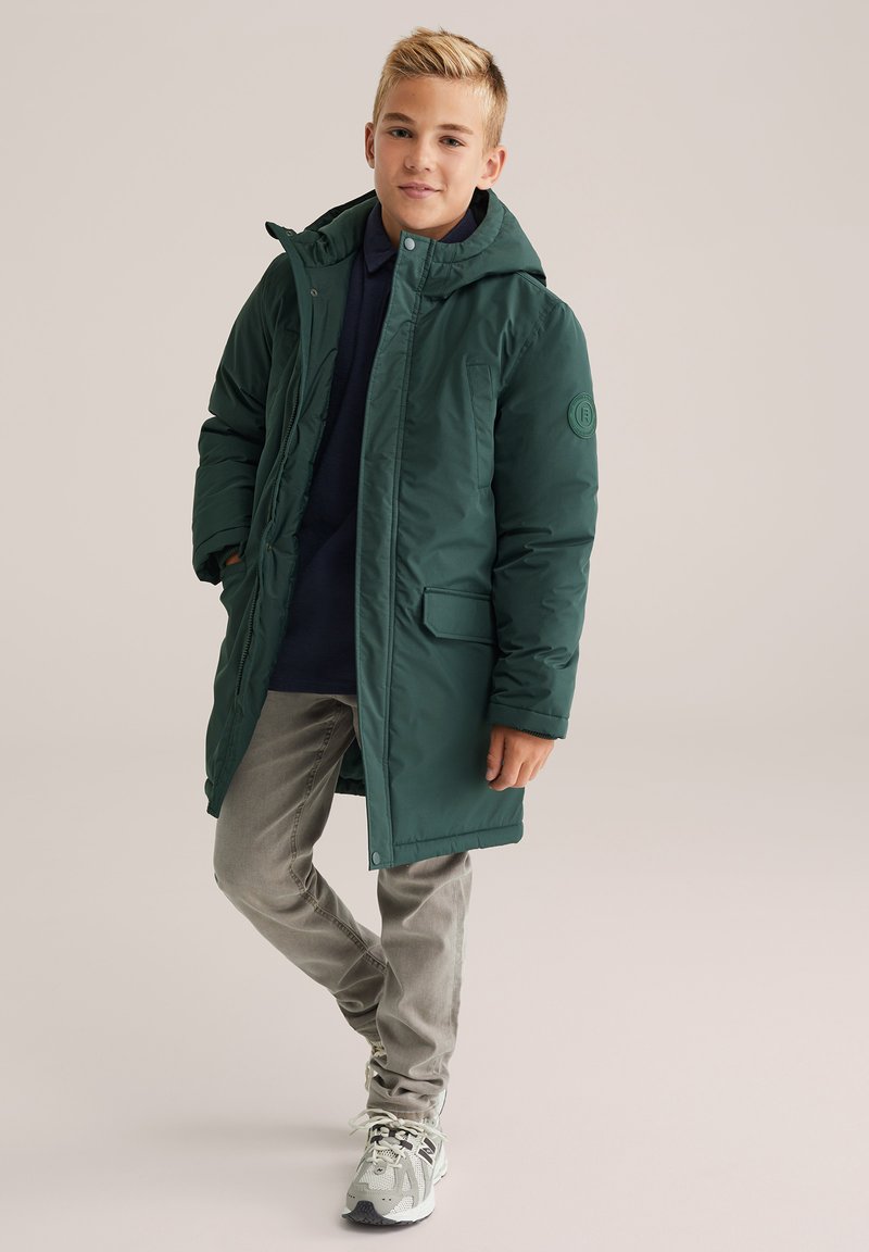 Jauns zēns stāv, ģērbies tumši zaļā kapuces ziemas mētelī, tumši zili krekls, bēšas bikses un pelēki sporta apavi, pret vienkāršu fonu.