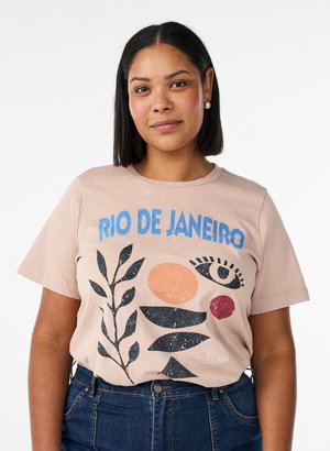 Kvinde med glat sort hår, der er iført perleøreringe, en beige t-shirt med teksten "RIO DE JANEIRO" og blå jeans mod en hvid baggrund.