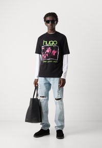 Svart t-skjorte med korte ermer og neongrønn og rosa grafisk design, brukt over en hvit skjorte med lange ermer. Lys blå revne jeans og svarte sko.