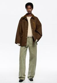 Hnedý kabát s kordovým golierom, obsahujúci cvokové gombíky, dve predné vrecká a voľný strih. Kombinovaný s svetlozelenými nohavicami so širokými nohavicami.