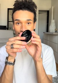 Schwarzer runder Behälter mit der Aufschrift "NYX MINERAL", der von einer Person in einem weißen Hemd gehalten wird. Das Produkt hat einen beigen Farbton. Heller Raum im Hintergrund.