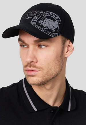 Mand med kort skæg iført en sort Lonsdale London kasket og sort skjorte med krave og grå stribe, kigger mod venstre.