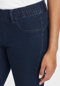 Jeans de mezclilla azul oscuro con cintura elástica, que presentan bolsillos laterales y una silueta ajustada. Textura suave con un sutil detalle de desvanecimiento.