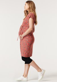 Vestido de maternidade em coral com padrão floral, mangas curtas, com laço ajustável na cintura, usado com leggings pretas e ténis brancos.