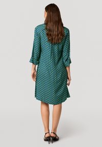 Langärmliges Kleid mit einem grün-blauen Muster, knielang, aus leichtem Stoff gefertigt, mit entspannter Passform und leichten Falten auf dem Rücken.