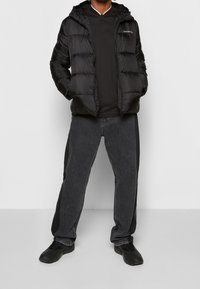 Giacca imbottita nera con cappuccio, dotata di chiusura con zip e cuciture orizzontali. Indossata sopra una polo nera e abbinata a jeans di denim scuro e scarpe nere.