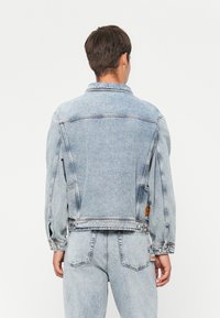 Hele sinine denim jakk klassikalise lõikega, millel on kaelus, pikad varrukad ja õmblustel õmmeldud detailsused.