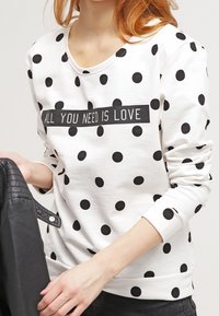 Biała bluza z czarnymi kropkami i czarnym napisem "ALL YOU NEED IS LOVE." Posiada okrągły dekolt i długie rękawy.
