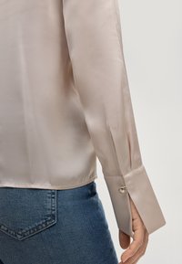 Lichtroze satijnen blouse met een soepele textuur, met een getailleerde pasvorm en een manchette versierd met een decoratieve knoop, gecombineerd met blauwe jeans.