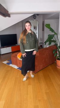 Ženska, ki stoji v notranjosti, oblečena v zeleno jakno, črno krilo in bele balerinke, blizu rjave zofe in velike rastline v loncu na lesenem tlaku.