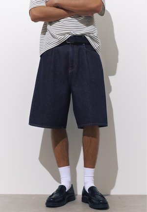 Homme les bras croisés, portant une chemise rayée, un short en jean foncé, des chaussettes blanches et des mocassins noirs, debout contre un mur blanc.