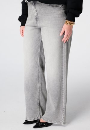 Femme portant un jean gris clair à jambes larges, des chaussures noires à bouts pointus, un haut noir et des bracelets dorés au poignet droit.