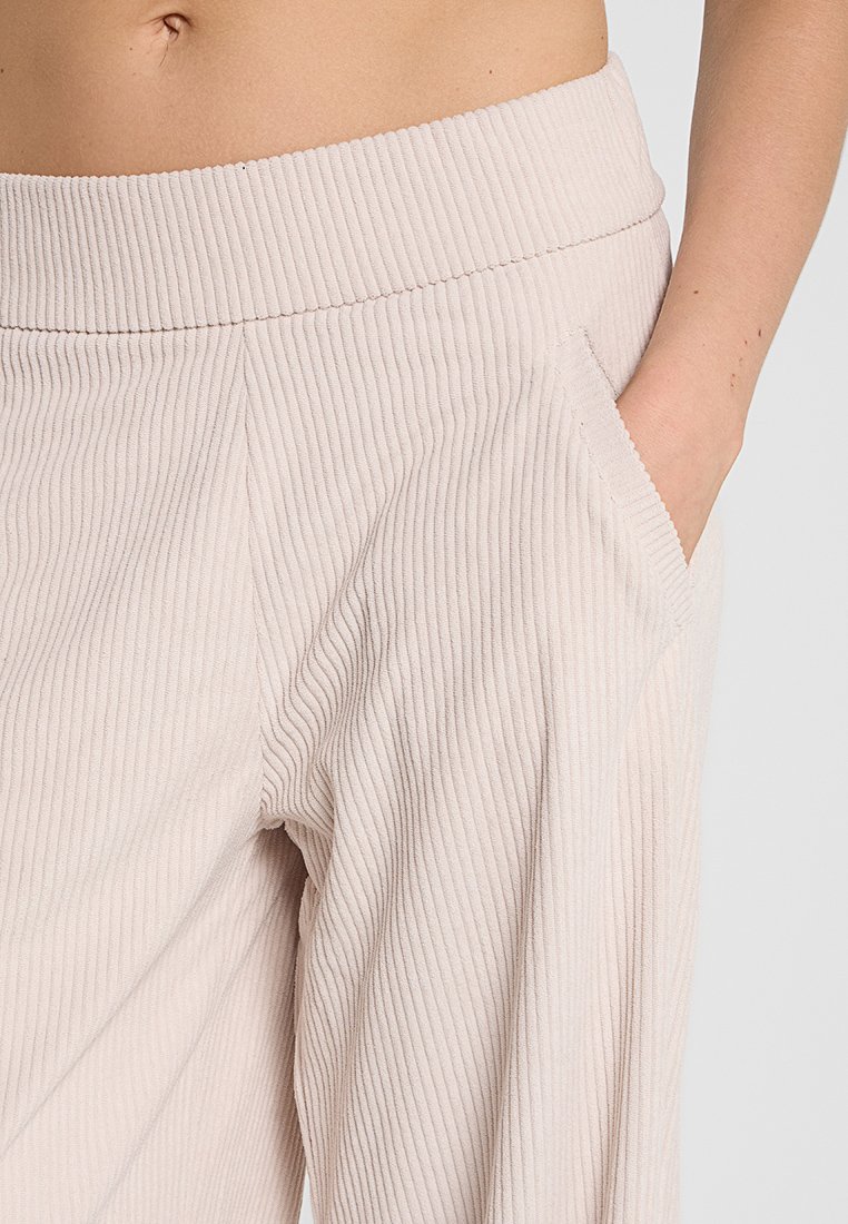Persona che indossa pantaloni a coste di colore beige chiaro con una mano in tasca, mostrando una parte del ventre e del braccio su uno sfondo chiaro.