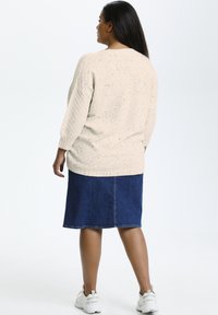 Beige textuurgebreide trui met kleurrijke spikkels, gecombineerd met een blauwe denim rok. Model draagt lichtgekleurde sportschoenen.