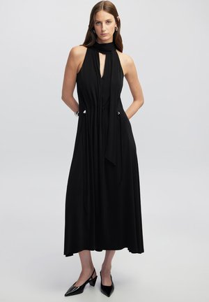 Robe noire sans manches avec un col haut, un décolleté en trou de serrure à l'avant et des liens sur les côtés. Tissu lisse, silhouette fluide et accents argentés près des liens.