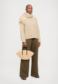 Pulover bej tricotat cu un model texturat, asortat cu pantaloni largi de culoare verde măsliniu. Poartă o geantă din nuiele, bej, cu mânere negre.