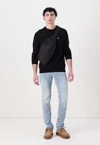 Zwarte schoudertas met een gladde textuur, ritscompartimenten en een verstelbare band, gedragen over het lichaam. Model in casual kleding.