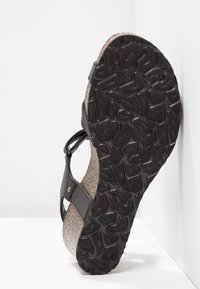 Semelle noire avec un motif irrégulier sur une sandale compensée en liège avec des brides noires, positionnée verticalement contre un fond blanc d’angle.
