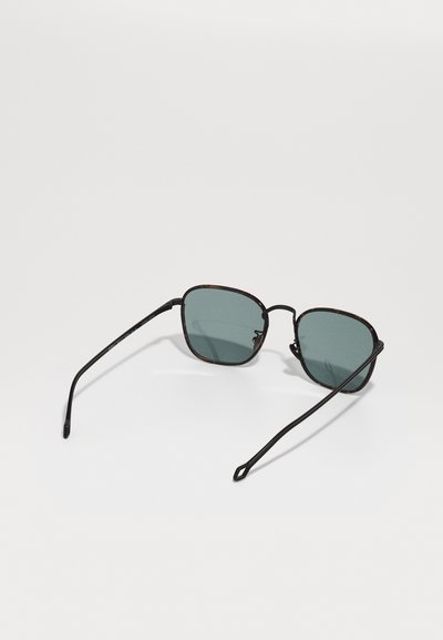 Ochelari de soare cu o formă rectangulară, cadru din metal negru și lentile verzi. Brațele sunt subțiri cu un detaliu geometric la capăt.