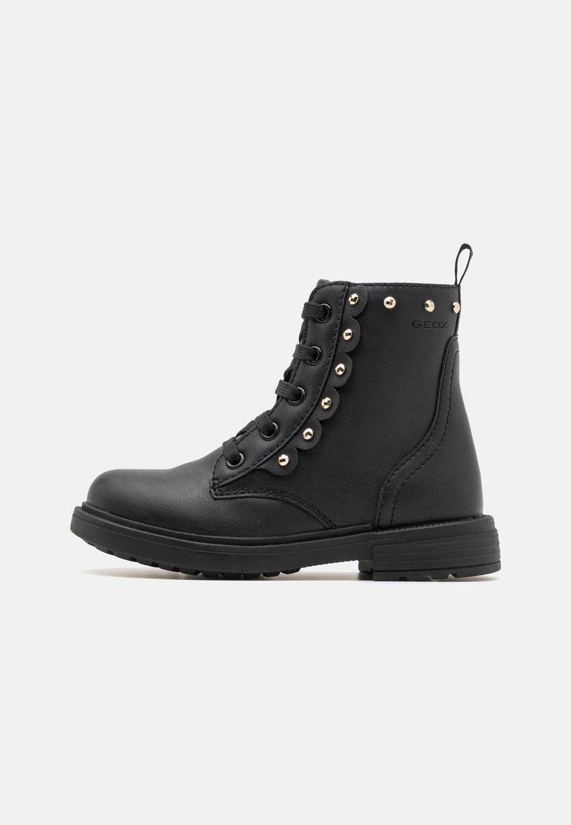 Bottine noire à lacets avec des clous en métal ronds le long du côté et un détail bord festonné, semelle plate et languette à l’arrière.