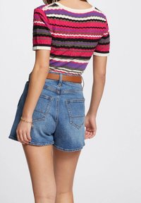 Kvinna iförd en flerfärgad randig kortärmad topp instoppad i högmidjade blå denimshorts med ett brunt bälte, sedd bakifrån.