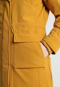 Veste jaune dotée d'une texture lisse, d'une fermeture éclair sur le devant et d'une grande poche latérale, avec un détail de manche à revers et une coupe droite.