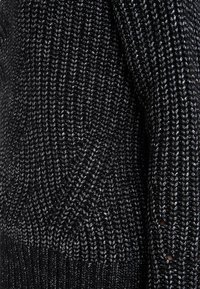 Svart stickad tröja med ett strukturerat mönster och metalltrådsdetaljer, med ribbad nederkant och detaljerad sömnad.