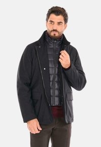 Giacca nera con zip frontale, colletto alla coreana e due tasche, sovrapposta a un gilet imbottito scuro. Texture liscia e vestibilità sartoriale.