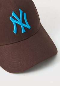 Hnedá baseballová čiapka s ohnutým vizírom a modrou vyšívanou ikonou so štylizovaným "NY" na prednej strane. Odolný materiál s textúrovaným povrchom.
